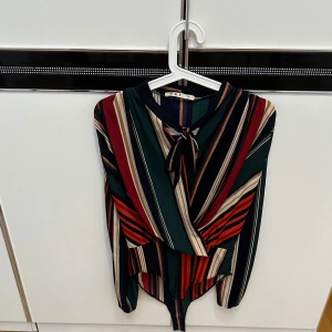 Randig blus med knyt från V&F - Säljer en färgstark randig blus från V&F i storlek S/M. Blusen/body har långa ärmar, knyt vid halsen och cool asymmetrisk nederkant. Färgerna är mörkgrön, vinröd, beige, marinblå och orange.