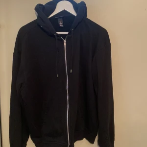 Svart hoodie med dragkedja från H&M - Svart hoodie från H&M i loose fit med dragkedja framtill och huva med snörning. Tillverkad i mjuk bomullsmix, perfekt för chill dagar. Långa ärmar och klassisk look som funkar till det mesta.