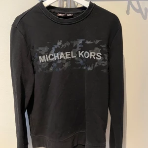 Svart Michael Kors sweatshirt M  - Svart sweatshirt från Michael Kors i bomull med coolt camo-tryck och stor logga över bröstet. Tröjan har rund hals, långa ärmar och en relaxed passform. Perfekt för dig som gillar streetstyle och vill ha något som sticker ut.