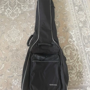 Svart gitarrväska från GEWA - Säljer ett svart gitarrfodral från GEWA. Fodralet har två ficka framtill med dragkedja och bärhandtag på sidan. Det finns även justerbara axelremmar på baksidan för att bära fodralet på ryggen. 