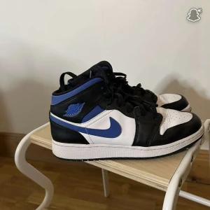 Nike Air Jordan 1 Mid svart/blå/vit - Säljer ett par Nike Air Jordan 1 Mid med svart, blå och vit färgkombination. Klassisk siluett med snörning, rund tå och vit sula. Ovandel i skinn och syntet, blå swoosh och detaljer. Perfekta för dig som gillar streetstyle och sneakers med ikonisk look.