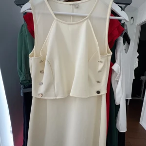 Krämvit ärmlös klänning med knappar - Snygg krämvit klänning med ärmlös design och dekorativa knappar på sidorna. Klänningen har en rak passform och är tillverkad i ett mjukt, slätt material. Perfekt för dig som gillar stilrena och enkla plagg med en twist.