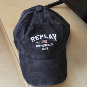 Replay svart keps NYC 1975 - Svart keps från Replay med böjd skärm och broderad text 'REPLAY NEW YORK CITY 1975' samt en liten amerikansk flagga framtill. Tillverkad i mjuk bomull med klassisk form och justerbar passform bak. Perfekt för dig som gillar streetstyle och snygga detaljer.