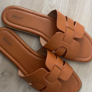 Bruna sandaler från Nelly - Snygga bruna sandaler från Nelly med flätad design och fyrkantig tå. Gjorda i syntetmaterial med platt sula, perfekta för varma dagar. 