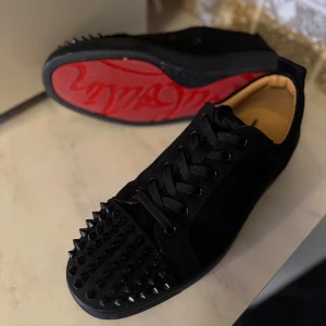 Christian Louboutin svarta sneakers med nitar - Svarta sneakers från Christian Louboutin i mocka och skinn med ikonisk röd sula. Tåpartiet är täckt av svarta nitar för en edgy look. Skorna har snörning och rund tå, perfekt för dig som vill sticka ut med lyxig streetstyle.