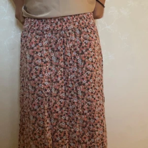 Blommig långkjol i rosa och lila - Supersöt långkjol med blommigt mönster i rosa, lila och vita toner. Kjolen har en avslappnad passform med resår i midjan och är gjord i ett lätt och luftigt material, perfekt för varmare dagar. Den ger en härlig boho-vibe och är enkel att styla med en enkel topp.