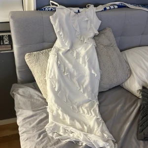 Vit långklänning med volanger - Säljer en elegant vit långklänning med tunna axelband och snygga volangdetaljer som går diagonalt över hela klänningen. Materialet är lätt och ger ett flowigt intryck, perfekt för dig som vill sticka ut med en romantisk look. Passar dig som gillar stilrena och feminina plagg.
