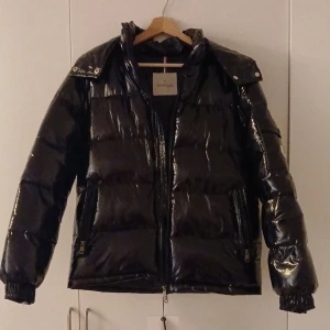 Svart glansig dunjacka från Moncler - Säljer en svart, glansig dunjacka från Moncler med klassisk pufferstil. Jackan har huva, dragkedja framtill och Moncler-logga på ärmen. Fylld med dun och har långa ärmar samt praktiska fickor med dragkedja. Perfekt för kalla dagar när du vill hålla stilen.