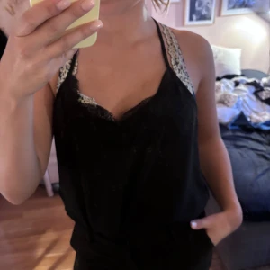 Svart linnetopp med spets från ONLY - Snygg svart linnetopp från ONLY med tunna axelband och v-ringad spetskant framtill. Toppen är ärmlös och har en mjuk, flowy passform som känns lätt och luftig. Perfekt att styla med jeans eller kjol för en trendig look.