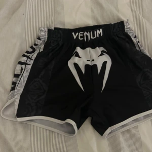 Svarta Venum träningsshorts M - Grymma svarta träningsshorts från Venum med vit logga och coolt mönster i grått och svart på sidorna. Shortsen har elastisk midja och snörning för perfekt passform. Perfekta för dig som kör kampsport eller annan intensiv träning.