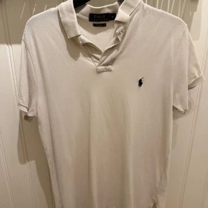 Vit pikétröja från Polo Ralph Lauren - Snygg vit pikétröja från Polo Ralph Lauren med klassisk krage och korta ärmar. Tröjan har den ikoniska broderade loggan på bröstet och är gjord i mjuk bomull. Perfekt för en clean och stilren look.