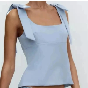 Ljusblå linnetopp med rosettband - Superfin ljusblå linnetopp med breda axelband som knyts i stora rosetter på axlarna. Toppen har en fyrkantig urringning och är lätt utsvängd nedtill. Perfekt för dig som gillar en söt och trendig look på sommaren.