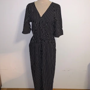 Svartvit randig jumpsuit Monki - Svart jumpsuit från Monki med vita vertikala ränder. Klänningen har v-ringning, trekvartsärm och resår i midjan för en snygg passform. Tyvärr saknar den en knapp men inget som märks