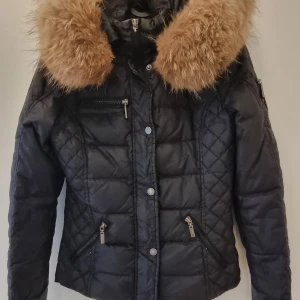 Svart pufferjacka Rockandblue med päls - Säljer en svart pufferjacka från Rockandblue med snyggt quiltat mönster och stor brun päls på huvan. Jackan har flera dragkedjor, cool patch på ärmen och sitter tajt och figurnära. Perfekt för kalla dagar när du vill vara både varm och stilren. Finns en lagad skada på ärmen. 