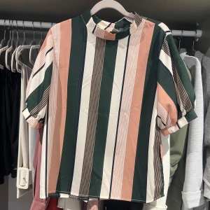 Randig blus med ståkrage - Snygg blus med breda vertikala ränder i grönt, vitt, rosa och beige. Modellen har korta ärmar, rak passform och en liten ståkrage med knapp i nacken. Perfekt för dig som gillar färg och grafiska mönster. Strl S/M