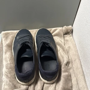 Svarta sneakers från Axel Arigato - Stilrena svarta sneakers från Axel Arigato med ovandel i skinn och detaljer i mocka vid hälen. Klassisk låg modell med snörning och vit platt sula. Diskret guldtext på sidan ger en clean look. Perfekta för dig som gillar minimalistisk stil.