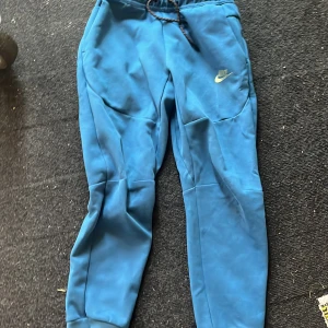 Blå mjukisbyxor från Nike - Snygga blå mjukisbyxor från Nike med klassisk logga på låret. Byxorna har dragsko i midjan och muddar vid bensluten för en skön passform. Perfekta för chill eller träning, tillverkade i mjukt material.