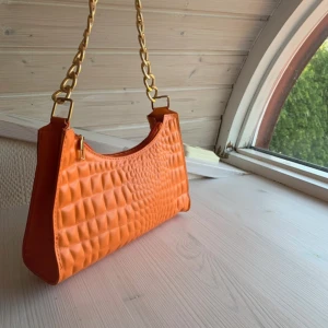 Orange quiltad axelväska med kedja - Snygg orange axelväska med quiltad struktur och guldfärgad kedja som axelrem. Väskan har en modern form och stängs med dragkedja upptill. Perfekt för att addera färg till din outfit och rymmer det viktigaste du behöver ha med dig.