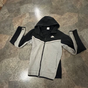 Nike hoodie svart och grå XS - Nike hoodie i svart och grått med dragkedja och huva. Snygg vit detalj över bröstet och klassisk Nike-logga. Långärmad och med bekväm passform, perfekt för chill eller träning. Tillverkad i mjukt material som känns skönt mot huden.