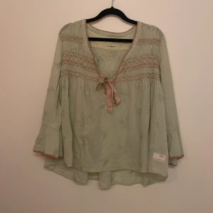 Ljusgrön blus Odd Molly broderad - Supersöt ljusgrön blus från Odd Molly med broderade klöver blommor och rosa detaljer. Blusen har v-ringning med rosett, vida ärmar och smockade partier framtill och baktill. Luftigt material och romantisk känsla, perfekt för vår och sommar.