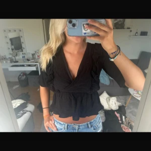 Svart omlottblus med volangdetaljer - Säljer en svart omlottblus med volangdetaljer och trekvartsärm. Blusen har en djup v-ringning och är lätt croppad med rynkad midja. Perfekt att styla med jeans eller kjol för en trendig look.