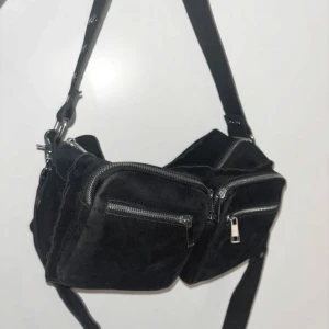 Svart axelväska i mocka från Noella - svart axelväska från Noella med två stora fack framtill och silvriga dragkedjor. Väskan har bred axelrem med nitar och flera innerfack för extra förvaring.
