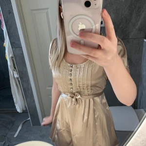 Champagnefärgad kort klänning med knappar - Snygg kort klänning i champagnefärg med glansigt material, knappar framtill och markerad midja med knytband. Puffiga korta ärmar och veckade detaljer på överdelen ger en elegant look. Perfekt för dig som vill sticka ut lite extra.