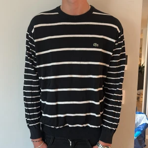 Randig svart tröja från Lacoste - Snygg svart tröja från Lacoste med vita horisontella ränder och klassisk rund hals. Tröjan har lång ärm och den ikoniska gröna krokodilloggan broderad på bröstet. Tillverkad i mjuk bomull, perfekt för en stilren och avslappnad look.