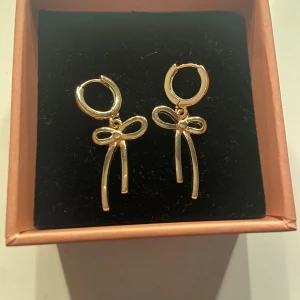 Guldiga örhängen med rosett - Supersöta örhängen i guld med hängande rosettformad detalj. Perfekta för att ge din look en extra touch av stil och charm. Säljs i en fin ask, redo att användas direkt!