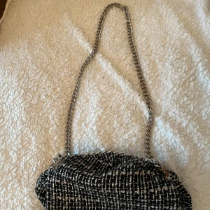 Svartvit axelväska med kedja - Säljer en snygg svartvit axelväska med rutigt mönster och grov kedja som axelrem. Väskan har en mjuk, textil yta och stängs med knäppe. Perfekt för dig som vill ha en trendig accessoar med lite edge.
