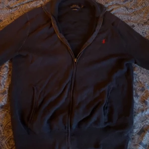 Mörkblå kofta från Polo Ralph Lauren - Säljer en mörkblå kofta från Polo Ralph Lauren i pima bomull. Tröjan har dragkedja framtill, ribbade muddar och två fickor. Klassisk röd broderad logga på bröstet. Perfekt för lager-på-lager under höst och vår.