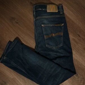 Nudie Jeans grim trim 30/32 - Mörkblå jeans från Nudie Jeans med klassisk femficksdesign och snygga orange sömmar på bakfickorna. Jeansen har en slim passform och är tillverkade i slitstarkt bomullsmaterial. Perfekta för dig som gillar en stilren och tidlös.