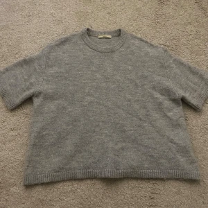 Grå stickad tröja från Gina Tricot - Mysig grå stickad tröja från Gina Tricot med rund halsringning och ribbade kanter. Tröjan har trekvarts ärmar och är perfekt för lager-på-lager-stil. Enkel att matcha med både jeans och kjol.