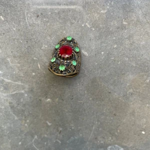 Vintage ring med röd och gröna stenar - Unik vintage ring i antikguld med en stor röd sten i mitten och flera små gröna stenar runtom. Ringen har ett öppet band och detaljerat filigranmönster som ger en cool retro vibe. Perfekt för dig som gillar statement-smycken och vill sticka ut.