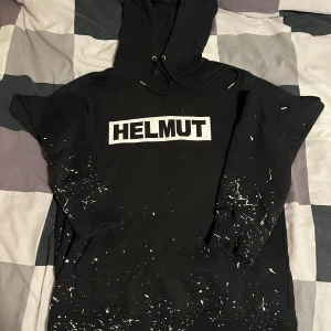Svart Helmut Lang hoodie med tryck - Svart hoodie från Helmut Lang med stor vit logga på bröstet. Tröjan har huva med snörning, känguruficka och coola vita färgstänk nertill. Perfekt för dig som gillar streetstyle och vill sticka ut lite extra. Den är i fint skick.