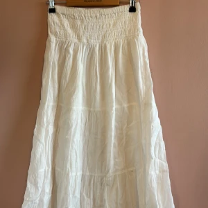 Vit långkjol från Gina Tricot - Luftig och flowy vit långkjol från Gina Tricot med smockad midja och flera volanger. Kjolen är tillverkad i ett tunt och mjukt tyg, perfekt för varma dagar. Enkel och stilren design som ger en chill och bohemisk vibe.