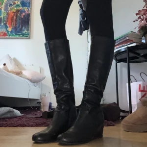 Svarta höga skinnstövlar - Snygga svarta stövlar i skinn med rund tå och låg klack. Klassisk modell som går upp till knäna och passar perfekt till både jeans och kjol. Stilrena och tidlösa boots som ger en cool vibe till din outfit.