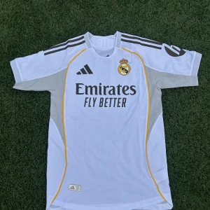 Real Madrid vit matchtröja Adidas S - Säljer en officiell Real Madrid fotbollströja från Adidas i storlek S. Tröjan är vit med grå och gula detaljer, har korta ärmar och är tillverkad i ventilerande polyester. Klassisk klubbmärke på bröstet och 'Emirates Fly Better' tryck på framsidan. Slim fit-modell.
