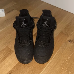 Svarta Nike Air Jordan 4 sneakers - Säljer ett par helsvarta Nike Air Jordan 4 sneakers med klassisk mesh och detaljer i mocka. Skorna har snörning, rund tå och den ikoniska Jumpman-loggan på plösen och hälen. Perfekta för dig som vill ha en stilren och sportig look.