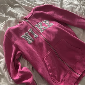 Vintage nike hoodie - Säljer en snygg rosa hoodie från Nike med dragkedja och stora vita bokstäver broderade på bröstet. Tröjan har två fickor framtill och är tillverkad i mjuk bomullsmix. Perfekt för chill dagar eller när du vill ha en sportig look.