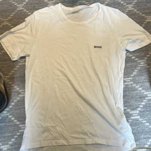 Vit t-shirt från BOSS, storlek L - Vit t-shirt från BOSS i storlek L. Klassisk rund hals och korta ärmar. Diskret BOSS-logga på bröstet. Mjuk och skön bomull som passar perfekt till jeans eller shorts. Enkel och stilren design som alltid funkar.