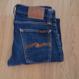 Blå jeans från Nudie Jeans - Säljer ett par klassiska blå jeans från Nudie Jeans med snygga kontrastsömmar och ikonisk bakficka med vågig brodyr. Jeansen har normal passform och är tillverkade i slitstarkt denimtyg av bomull. Storlek 27/32