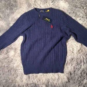 Blå stickad tröja från Polo Ralph Lauren - Mörkblå stickad tröja från Polo Ralph Lauren med klassisk rund hals och ribbade muddar. Tröjan har ett diskret kabelstickat mönster och den ikoniska röda loggan broderad på bröstet. Perfekt för dig som gillar stilren och tidlös design.