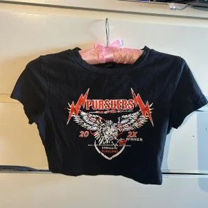 Svart croppad topp från SHEIN - Svart croppad topp från SHEIN med coolt tryck framtill i rött och vitt, med texten 'Pursuers' och en stor örn med utbredda vingar. Toppen har korta ärmar och rund hals. Perfekt för dig som gillar edgy och rockig stil.