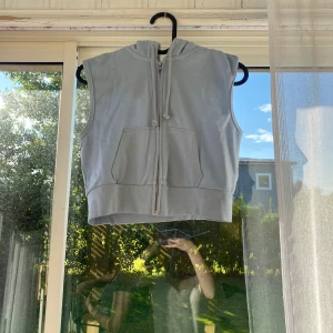 Ljusgrå croppad hoodietopp - Säljer en ljusgrå croppad hoodietopp med dragkedja framtill och stor ficka. Toppen är ärmlös och har en snygg huva med snören. Perfekt för chill dagar eller när du vill ha en sportig vibe. Mjuk bomullskänsla och loose passform.