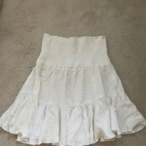 Vit volangkjol med smock Lager 157 - Supersöt vit kortkjol med bred smock i midjan och volang nertill. Kjolen har en luftig känsla och passar perfekt till varma dagar. Enkel att matcha med crop tops eller stickade tröjor. Använt 1 gång 💗