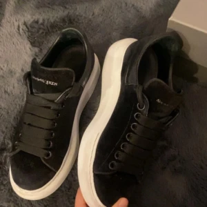 Alexander McQueen svarta sneakers - Svarta sneakers från Alexander McQueen med bred vit platt sula och klassisk snörning. Ovandelen är i mocka och skinn, med logga på plösen. Snygg chunky siluett som ger en cool vibe till outfiten.