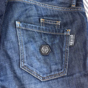 Philipp Plein straight cut jeans blå - Snygga blå jeans från Philipp Plein med straight cut passform. Jeansen har slitna detaljer, klassiska fem fickor och logotyp på bakfickan. Tuffa kontrastsömmar och knappgylf med märkets namn. Inga fel.