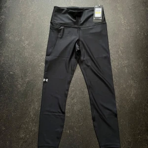 Svarta leggings från Under Armour - Svarta leggings från Under Armour med hög midja och diskret logga på benet. Tillverkade i stretchigt syntetmaterial som andas, perfekta för träning. Modellen har en tight passform och praktiska sidfickor för småsaker. Finns i stlk S M L