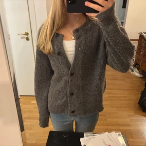 Mörkgrå stickad kofta med knappar - Mysig mörkgrå stickad kofta med rund hals och svarta knappar framtill. Jättefint skick och köpt i våras för 900kr.